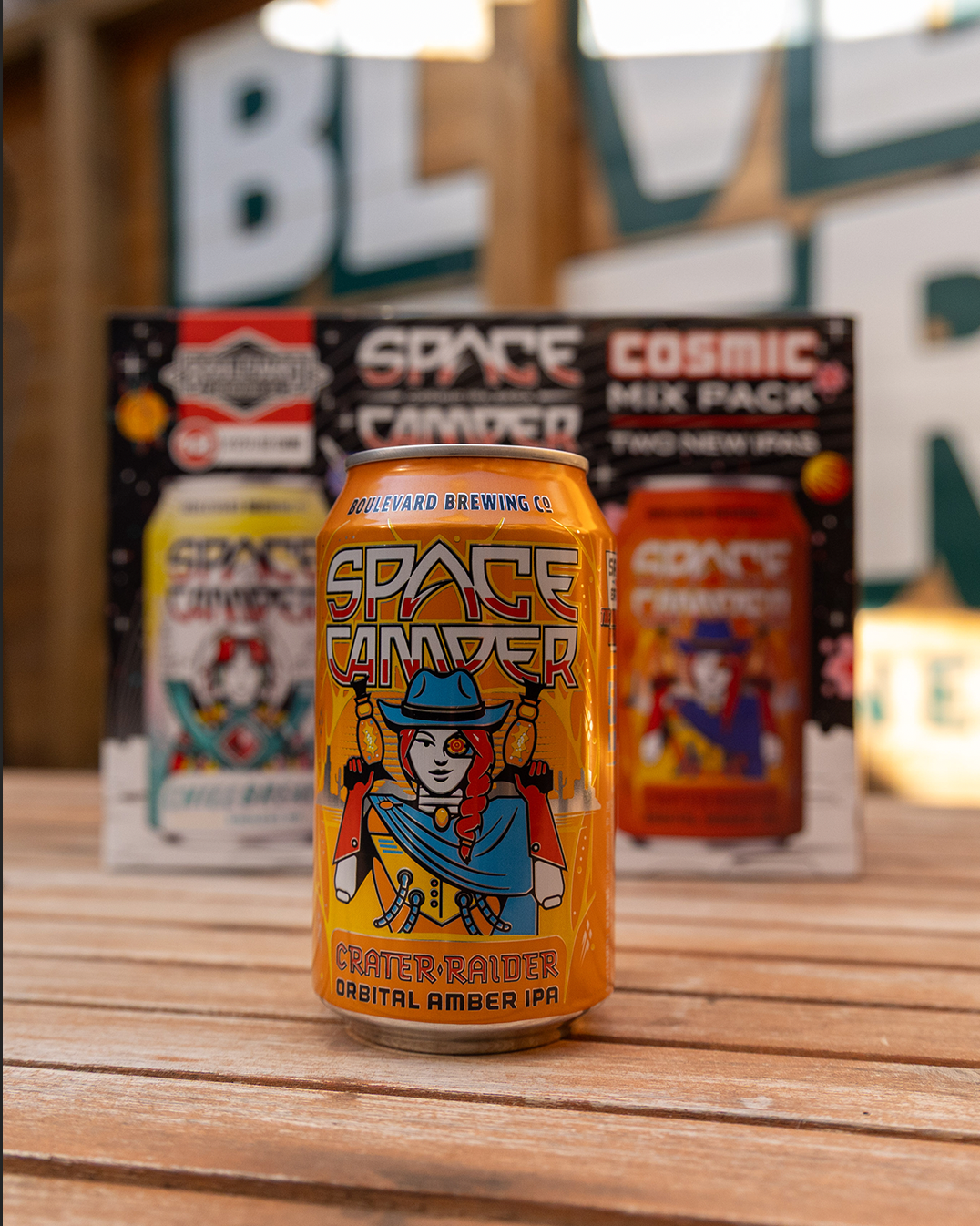 A can of Boulevard Brewing Co.'s Space Camper beer is on a wooden table. The artwork on the can features a futuristic astronaut. Blurred boxes of the Cosmic Mix Pack are visible in the background.